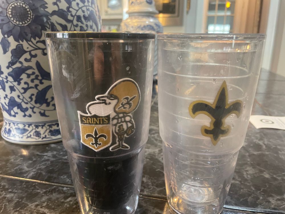 Two large New Orleans Saints Tervis tumblers, the one on the right should be clear but is cloudy from her handling it and has a black and gold fleur de lis on it. The one of the left has a black interior and white streaks all over it from her using it with her essential oils. It has Sir Saint on it with him holding a Saints crest.