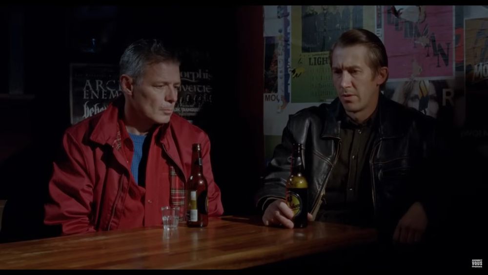 Deux personnages une bière à la main dans un bar, affiches de Metal derrière eux.