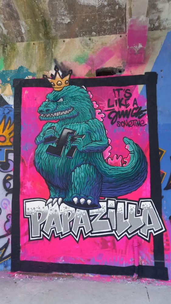 Graphe très coloré d'un Godzilla avec une couronne sur la tête, debout sur le nom "Papazilla" et brisant de ses pattes une croix gammée. Graphe signé Jungle (Uzerche, 2025) 