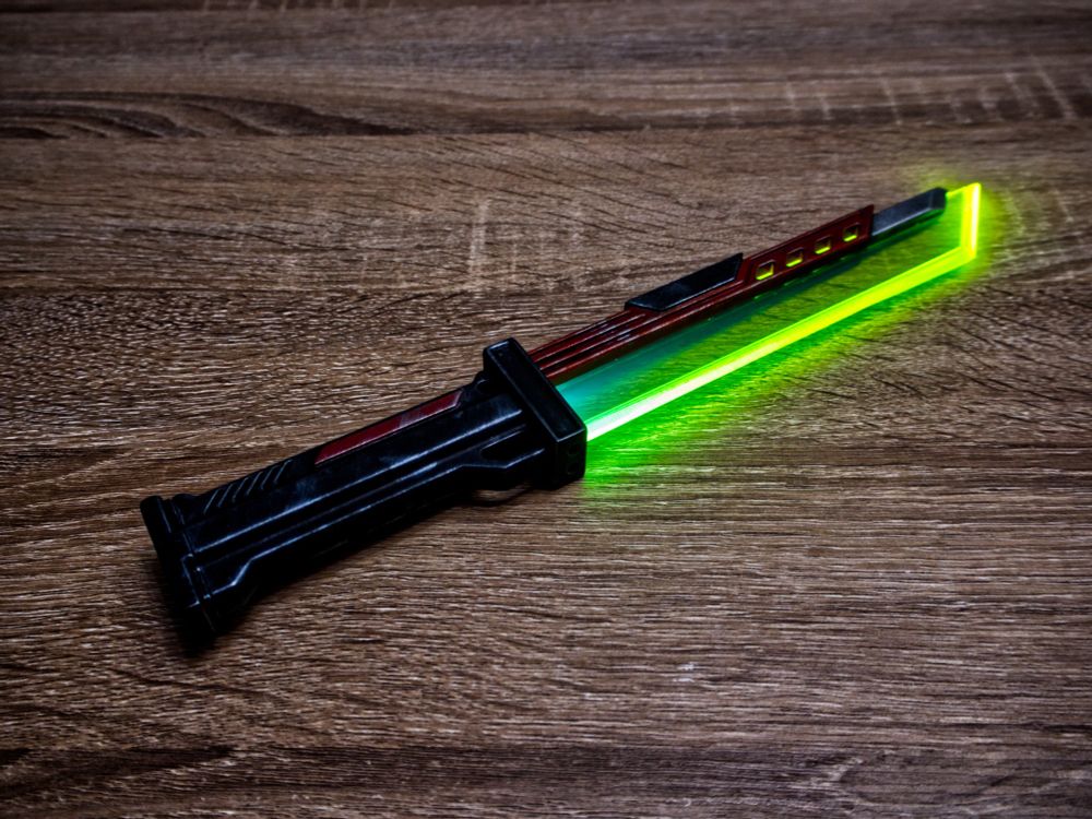 Picture of the 3D printed cyberpunk knife. The knife is finished and painted in black and red. It is weathered, with used metal effect.
The blade is in a green/yellow fluorescent acrylic. The light is on and the blade glows.
The knife if vertically layed on a wood table. The picture is taken from another angle.