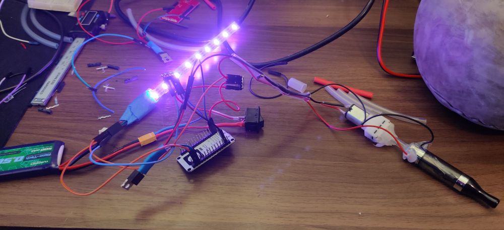 Montage électronique chaotique reliant des bandes de LEDs et une mini machine à fumée à un ESP8266. La machine à fumée est contrôlée par un MOSFET relié également à l'ESP. Un interrupteur permet d'allumer le circuit. Le tout est alimenté par une batterie 7.4v