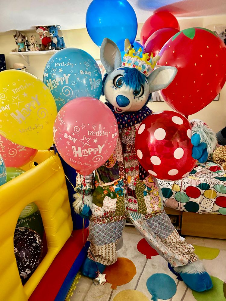 blue deer in a clown outfit, wearing an inflatable crown, next to a small bounce castle while holding helium birthday balloons 