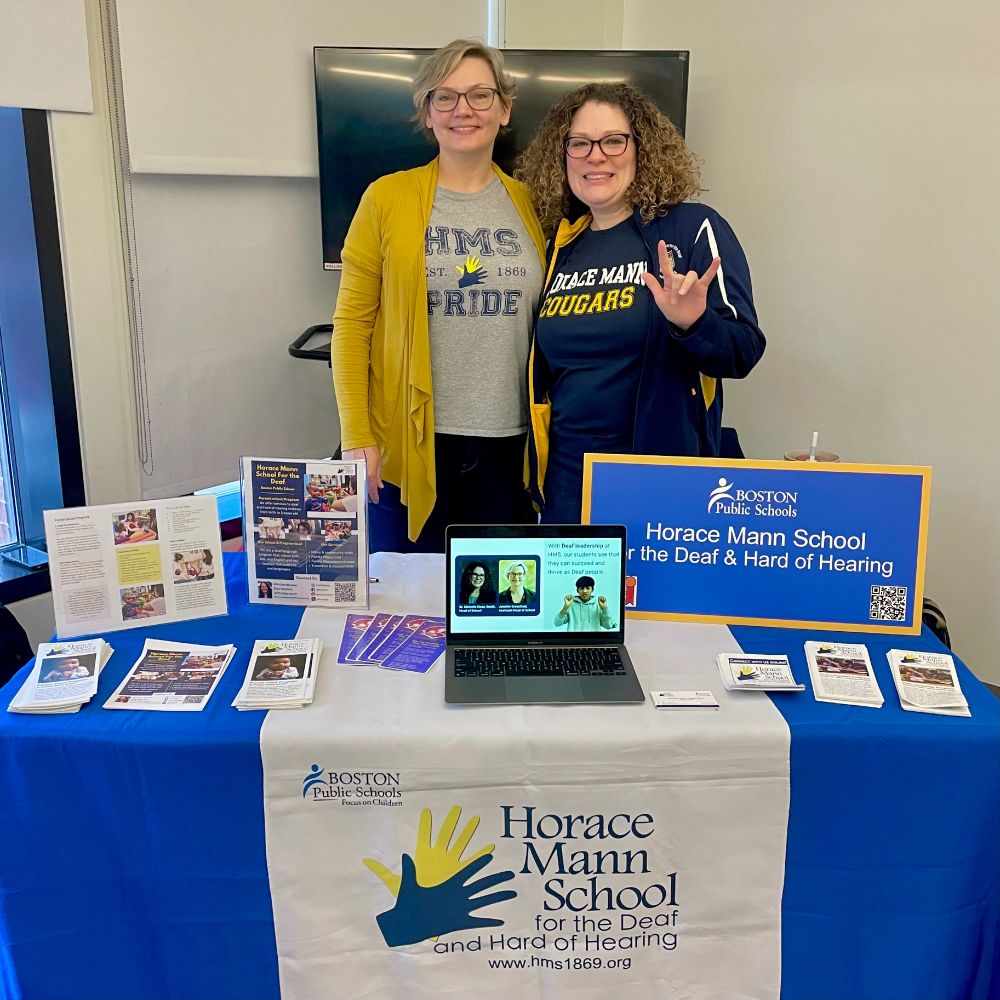 Photo shows two Heads of School from the Horace Mann School for the Deaf and Hard of Hearing standing in front of the HMS booth at the annual Boston Public Schools Showcase of Schools, smiling at the camera.