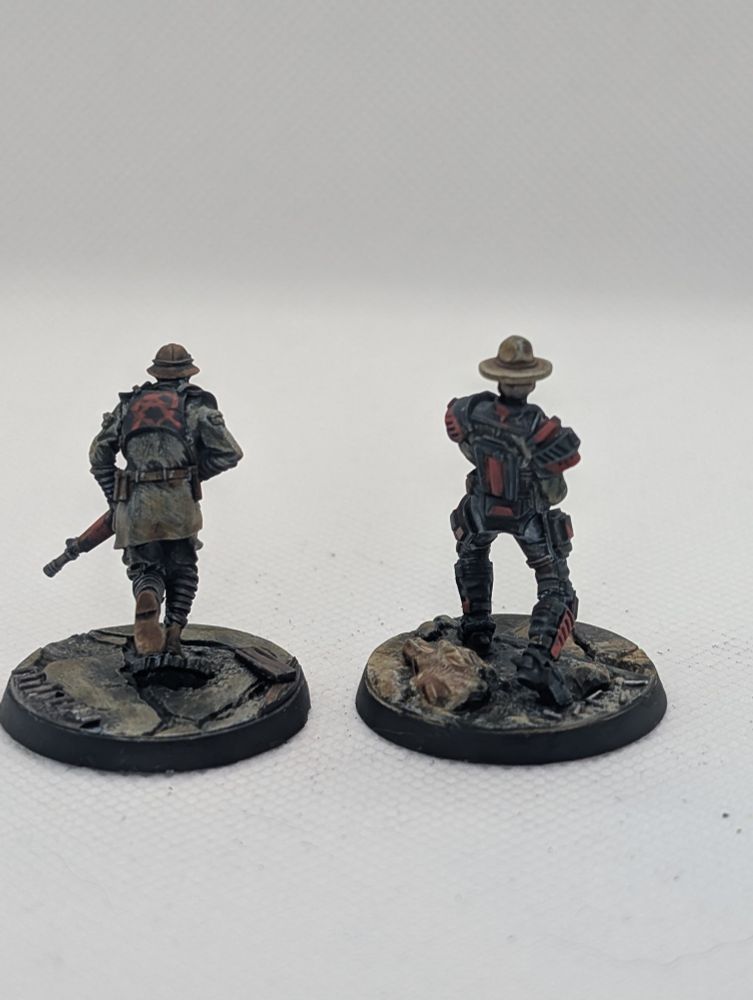 A pair of NCR minutes from the game fallout wasteland warfare, one is a trooper and one a ranger, they are painted in a dirty and grimy scheme with tan, leather and black as the main colours with red accents. The minis are pictured from behind, one has a crude anarchy symbol drawn across this armor.