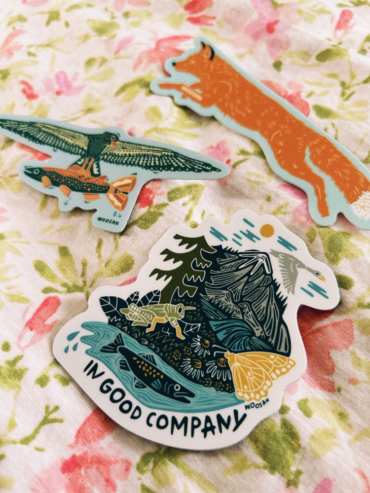 3 stickers on floral print bedding: a sticker with mountains and bugs and a fish in water that says “in good company”, another sticker of an osprey bird carrying a salmon in its claws, and the last sticker is a jumping fox