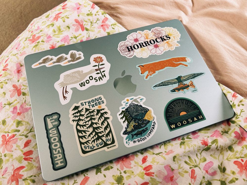 a blue macbook air on floral bedding; it’s covered in various stickers from a small local artist