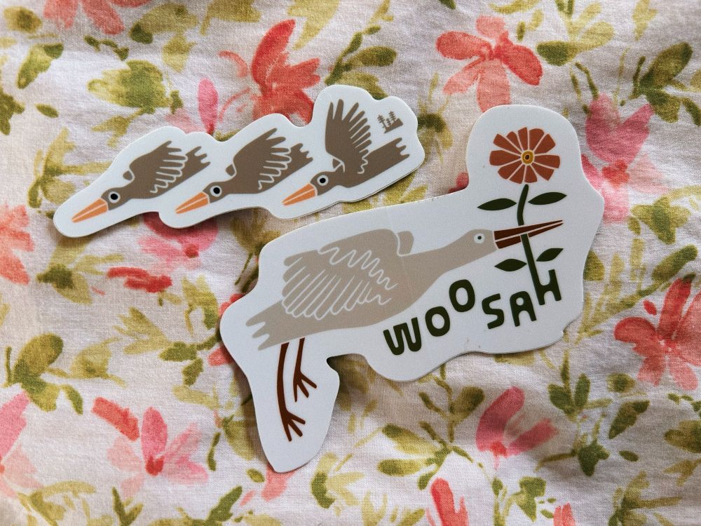 2 stickers on floral bedding: one is of three birds flying in a line, another is of a bird carrying a flower with the logo of the artist that says “woosah” 
