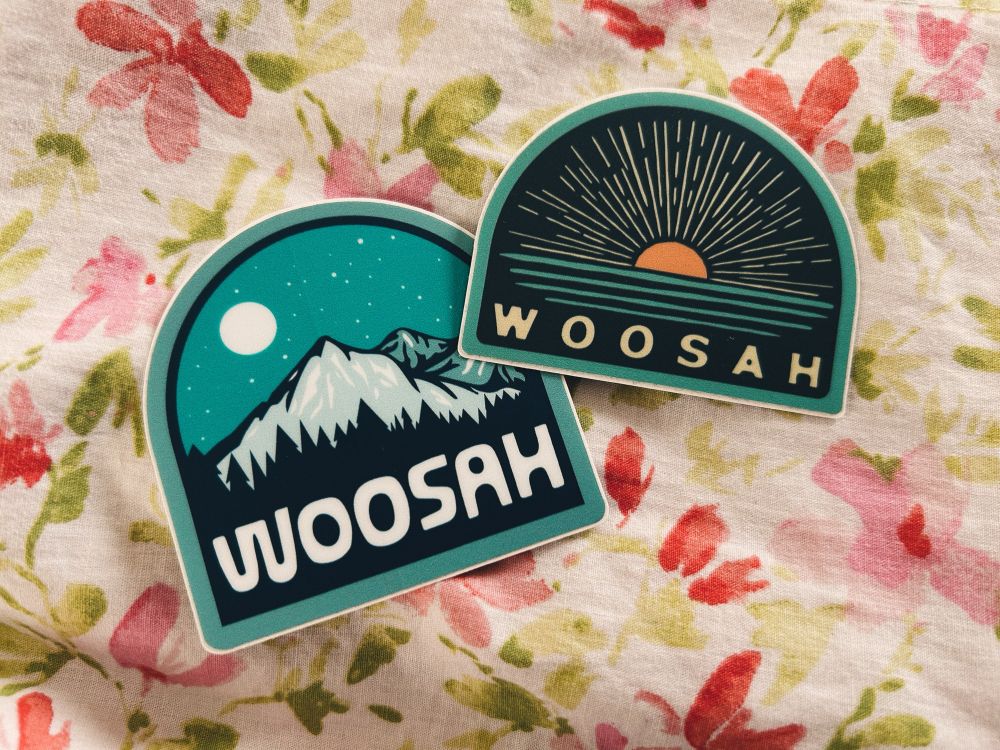 2 stickers on floral bedding: one is of a blue mountain with a foreground of pine trees and a sparkling night sky with the moon and the other is of a rising run over a lake; both with the artists logo underneath that says “woosah”