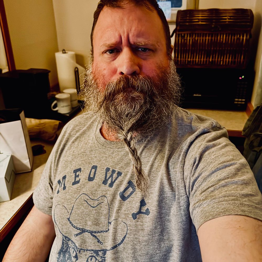 Bearish man with a braided beard wearing a grey “meowdy” t-shirt with a cat in a cowboy hat. 
