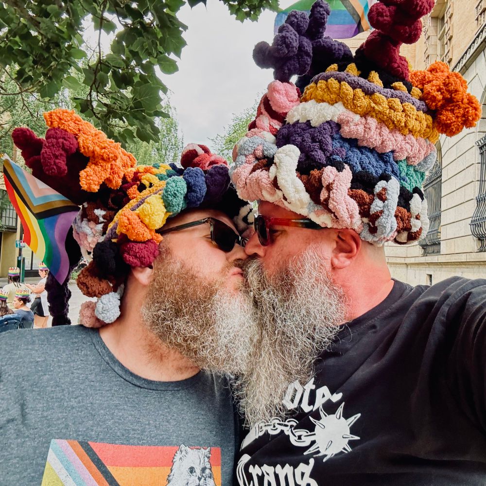 Bearish, bearded men in pride-colored yarn hats and sunglasses, kissing. 
