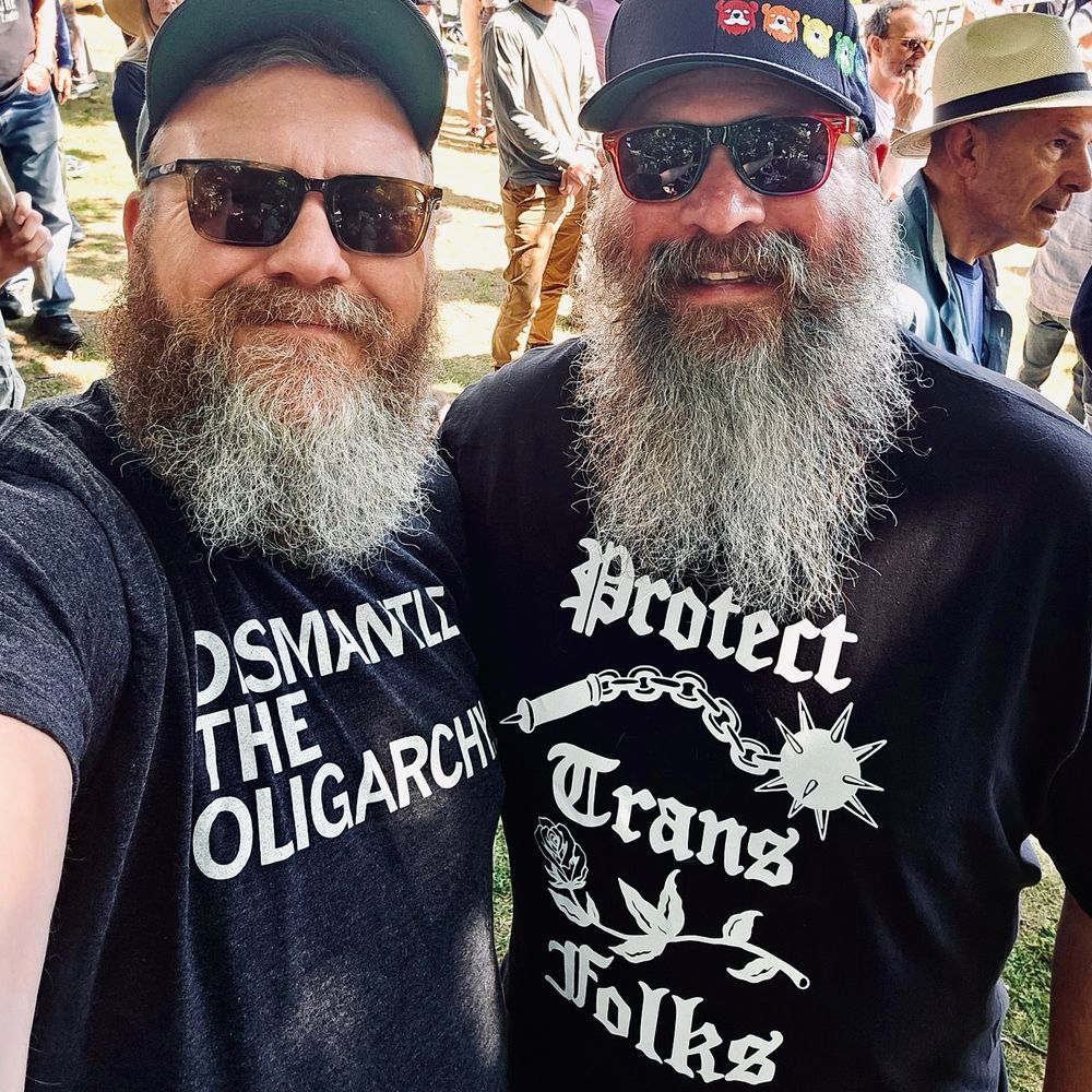 Two bearish men at a protest wearing shirts that say “dismantle the oligarchy” and “protect trans folks”. 