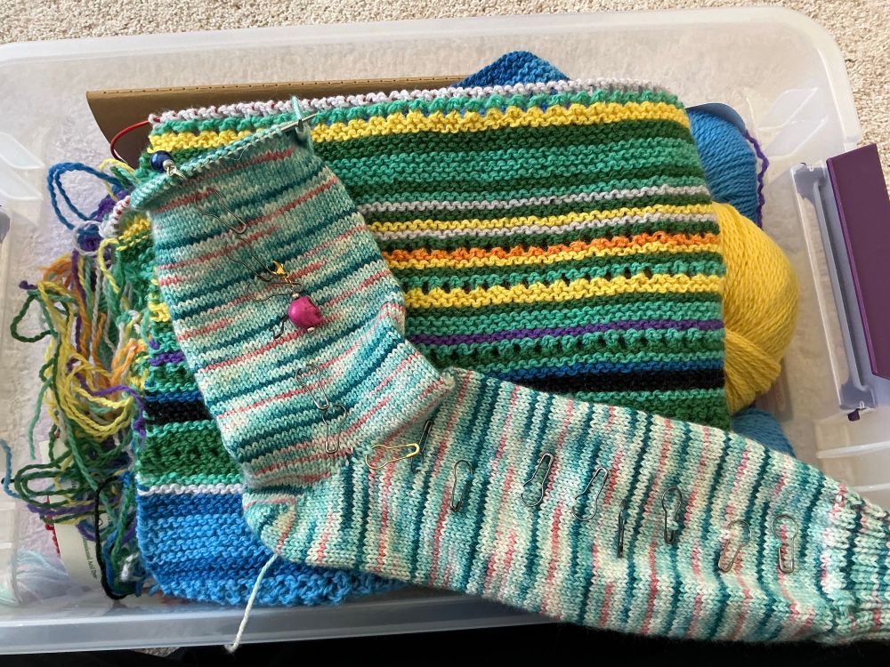 Knitting projects stacked together. On the bottom is a multi-colored, folded, and striped garter stitch wrap. On top is a plain sock nearly knit to the toe decrease in pinks and blues.