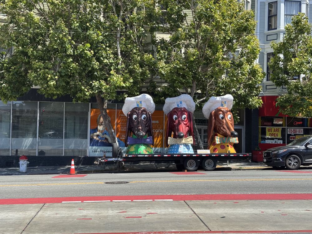 Three large sculpture that resemble a dog’s head. The dogs are wearing chef hats and outfits. They are the old mascots from the Doggie Diner chain that used to be in the city. These three sculptures are parked on a large wagon on Mission Street. 