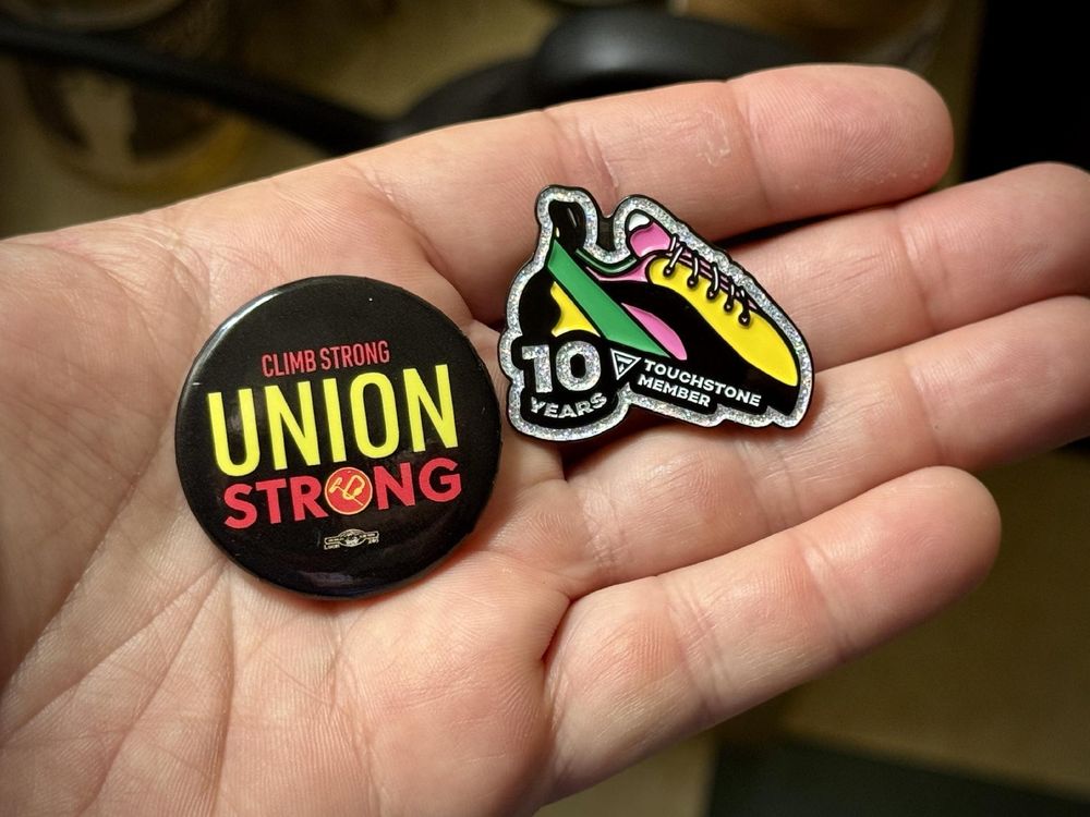 A hand holding a "Touchstone Workers United" button and a "10 Year Membership" enamel pin. The enamel pin is in the shape of a climbing shoe and was a gift for gym members who have been there longer than 10 years. 