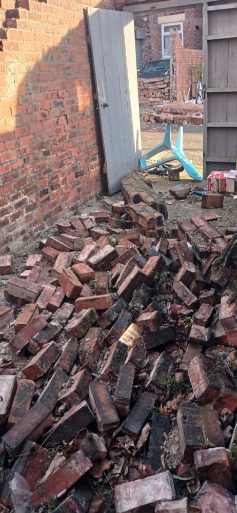 A large pile of bricks from a wall, lying in the foreground. Behind is an open gate which should be locked. An upturned plastic chair is in the gateway. Behind the chair are more fallen bricks from walls belonging to other houses.