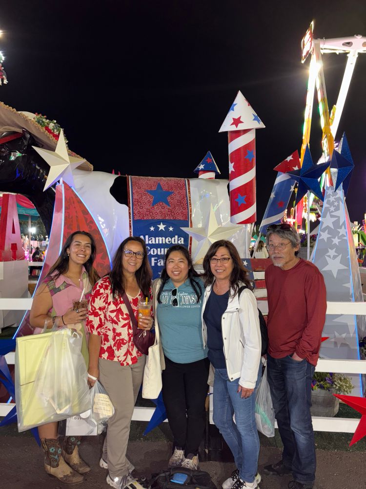 Photo of me and my family (4 Asian-American women, 1 Asian-American man) in front of a fake cow display that has a sign that says Alameda County Fair. The decorations around the display are red, white, and blue. 