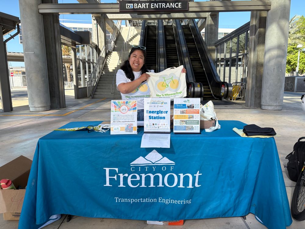 Photo of me holding up a Bike to Wherever Day canvas tote bag at an Energizer Station hosted by the City of Fremont, Transportation Engineering. The background is the stairs and escalators at a BART station