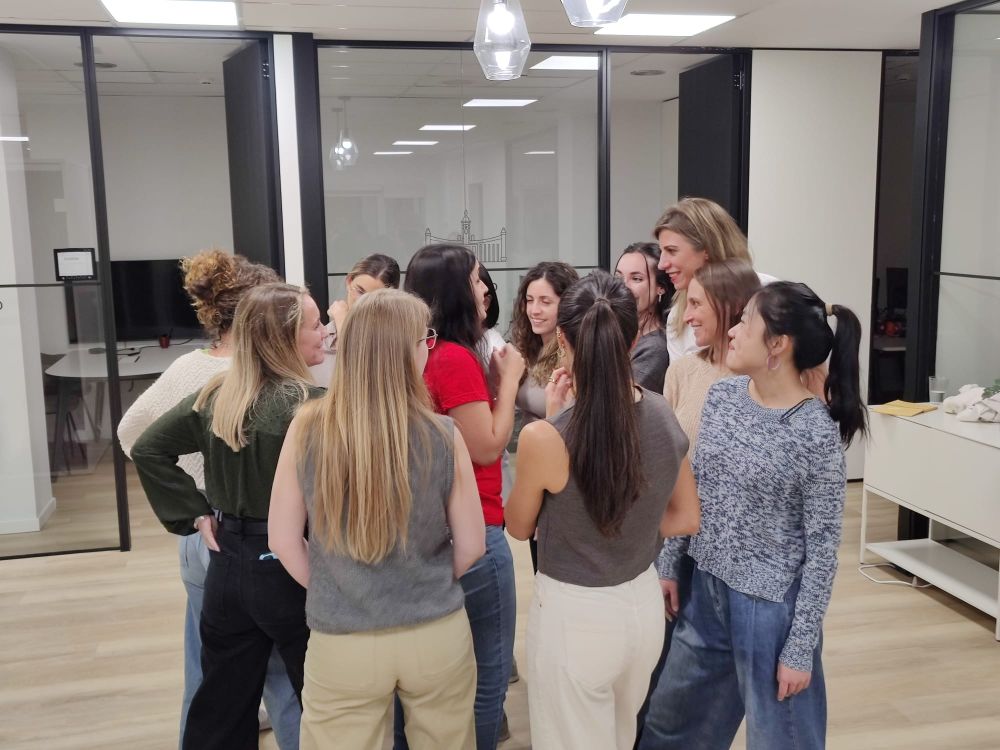 Foto de una de las dinámicas de la sesión, con María explicando el ejercicio mientras las participantes, de pie y formando un círculo estrecho a su alrededor, sonríen.