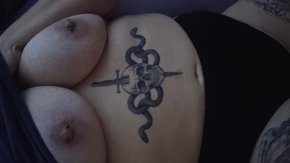 In bed,  shirt pulled up showing boobs and tummy. Tattoo and piercings visible 