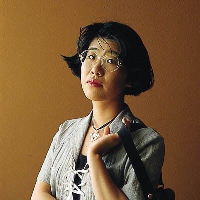 An early-career color photograph of young Banana Yoshimoto. She's facing away from the camera, but looking into the lens straight on with a wry expression. Her thick, black hair is bobbed close to her ears. She's wearing a gray-green lace-up dress or top over a black shirt and a ribbon choker with a large, translucent pendant. She's casually slinging a bag over her left shoulder using her left arm. Her thick, round, clear plastic glasses complete the look. The overall effect is of a woman with style and sass.