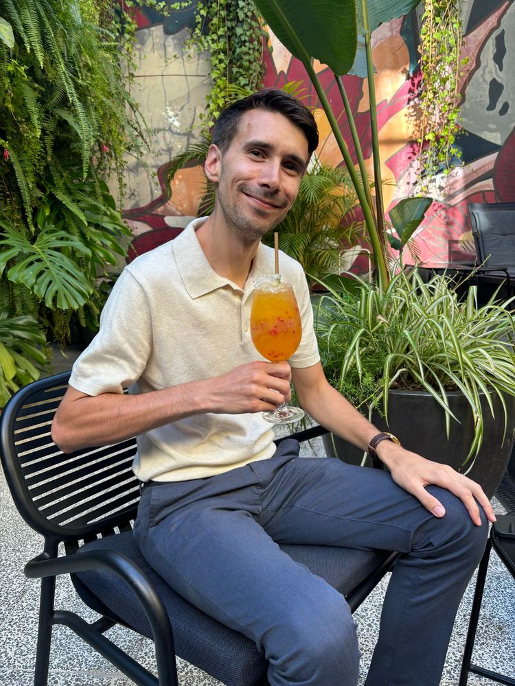 Austin sitting down, holding cocktail in front of a mural and plants 