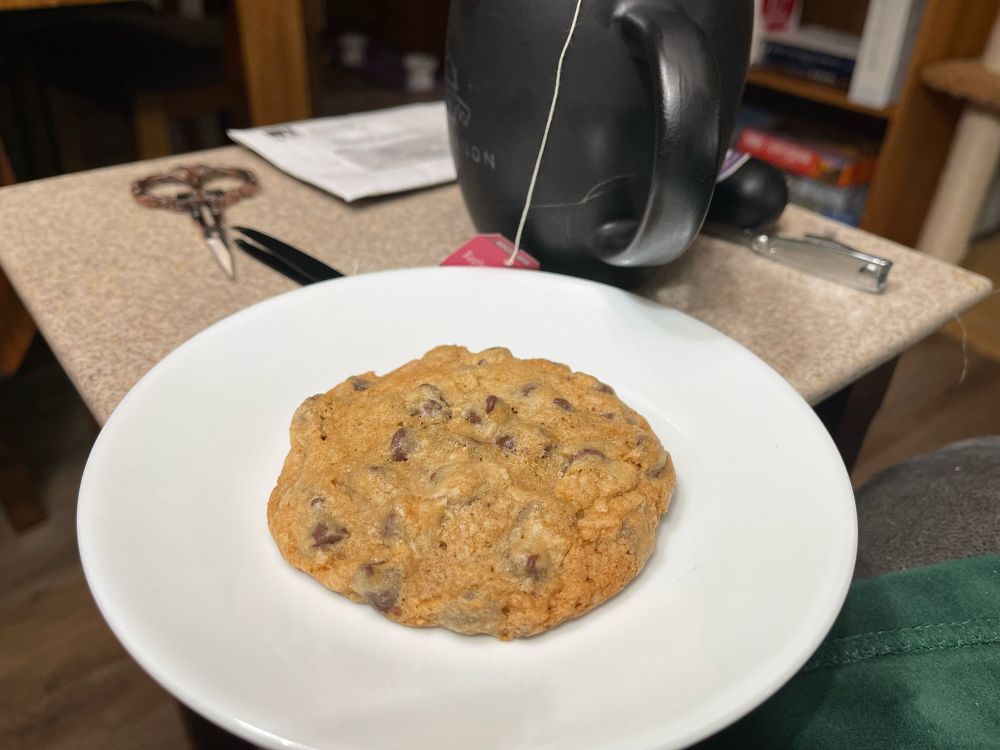 A chocolate chip cookie on a plate with a cup of tea.