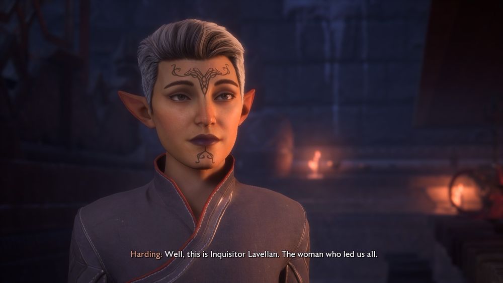 A pale elven woman with pixie-short grey hair, pointed ears, and dark lips. A curling-swirl tattoo sits over her forehead and down her nose, ending under her lips. She has a serious expression, and is dressed simply in a blue military tunic. The caption below her reads, “Well, this is Inquisitor Lavellan. The woman who led us all.”