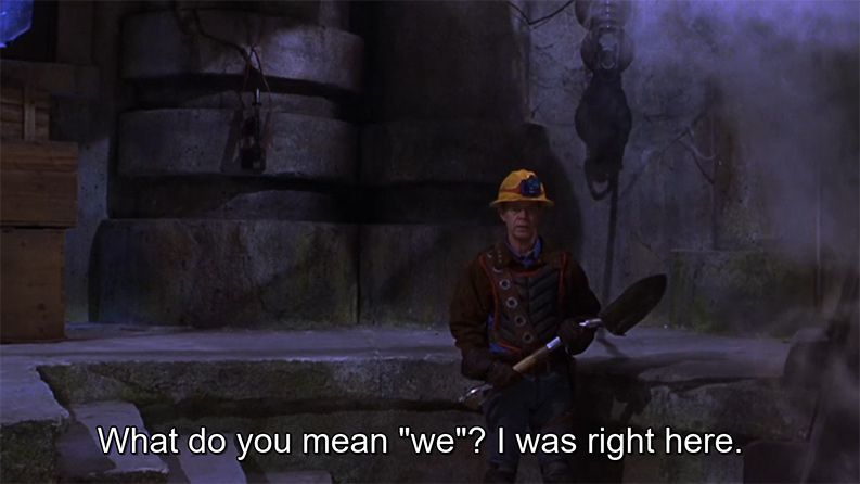 shot of actor William H. Macy wearing a yellow safety helmet with headlamp, a padded vest, leather jacket & gloves. he’s holding a shovel & standing a little to screen right. the background is a smoky disused concrete bunker. subtitle: “What do you mean ‘we’? I was right here.”