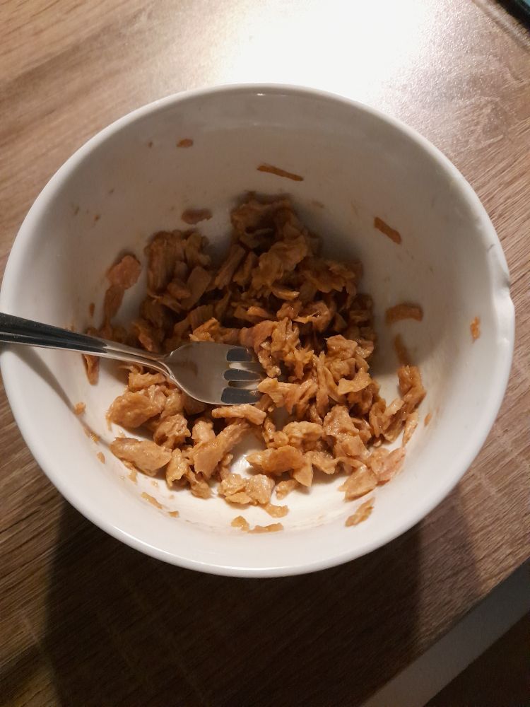 Angerührte Thunfisch Alternative in einer Porzellanschüssel. 
Das Bild ist von oben aufgenommen. 
Auf einer Küchenarbeitsfläche in Holzoptik steht eine weiße kleine Porzellanschale. Darin ist eine optisch eins zu eins aussehende Thunfisch Alternative, die gequollen ist. Links liegt eine Kuchengabel aus Edelstahl in der Schüssel. 