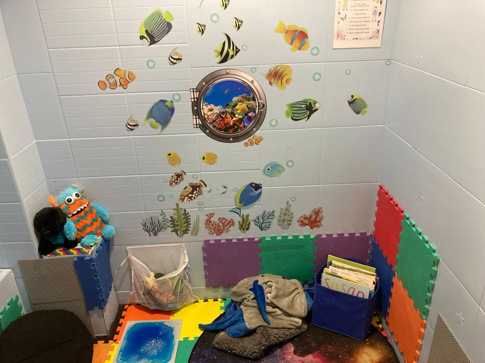 A sensory room with light blue walls, fish and bubble stickers, bean bags, a white bag of fidget toys and plushies, a book box and foam panels on the walls and floor. 