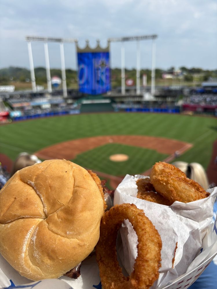 Kauffman Stadium panorama before an afternoon game. Sun, green grass, clouds and blue sly, with a Z-man barbeque sandwich an onion rings in the foreground. 