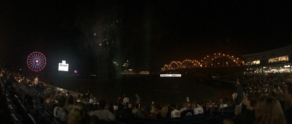 Post-game fireworks at a Quad Cities River Bandits game. Beyond left field is a ferris wheel lined with red and white lights, smoke from fireworks in centerfield, and a metal arch bridge outlined with white lights beyond right field. 