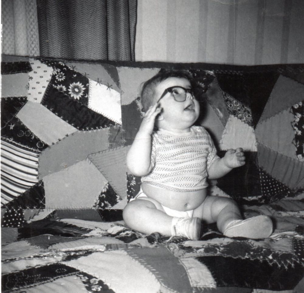 A 7-month-old baby (me) sitting on a sofa covered in a crazy patchwork quilt wearing a belly-revealing shirt, diaper and white walker baby shoes. I'm modeling my dad's dark-framed glasses, with one hand reaching toward them to either hold them on or take them off!