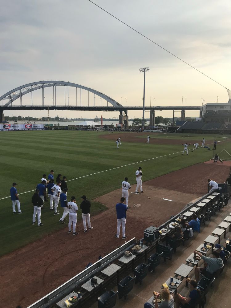 A group of pitchers watch another pitcher  warm up in the right field bullpen area before a Quad Cities River Bandits game. Modern Woodmen Park sits on the bank of the Mississippi River, which is in the background along with a metal arch bridge  over the water. 
