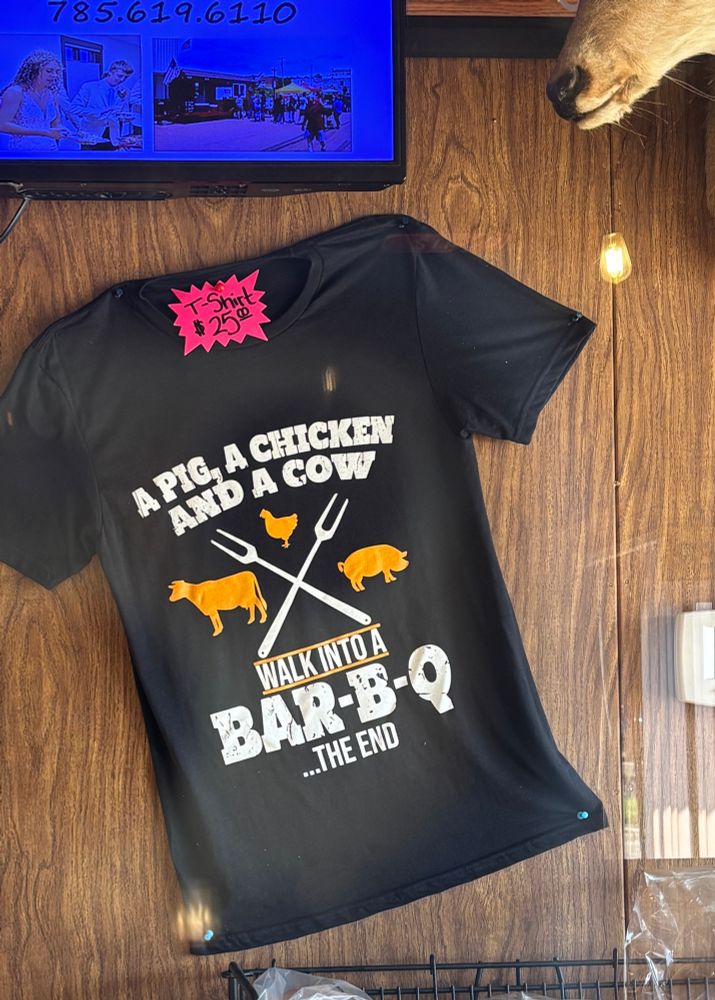 A black t-shirt with a logo of crossed grilling forks, with a cow, chicken and pig inside three of the quadrants. The logo above and below the image reads: “A PIG, A CHICKEN AND A COW WALK INTO A BAR-B-Q … THE END”