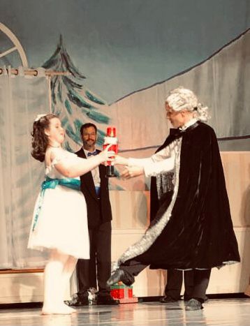 A scene from the Nutcracker. Clara in a knee-length white dress with a bright blue belt accepts a wooden nutcracker being handed to her by Herr Drosselmeyer, who is wearing a black velvet cape. 