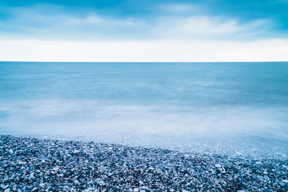 Una scena tranquilla di una spiaggia di ciottoli sotto un cielo nuvoloso. In primo piano, la spiaggia è ricoperta di piccoli sassi tondi di vari colori; sullo sfondo si vede il mare calmo che si fonde con l’orizzonte e il cielo azzurro con leggere sfumature grigie. L’atmosfera è serena e silenziosa.