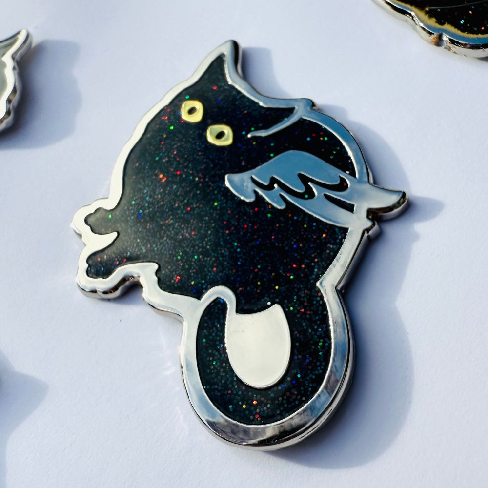 A photo of an enamel pin of a round cat with wings with black rainbow glitter enamel and dual-plated gold and silver metal.