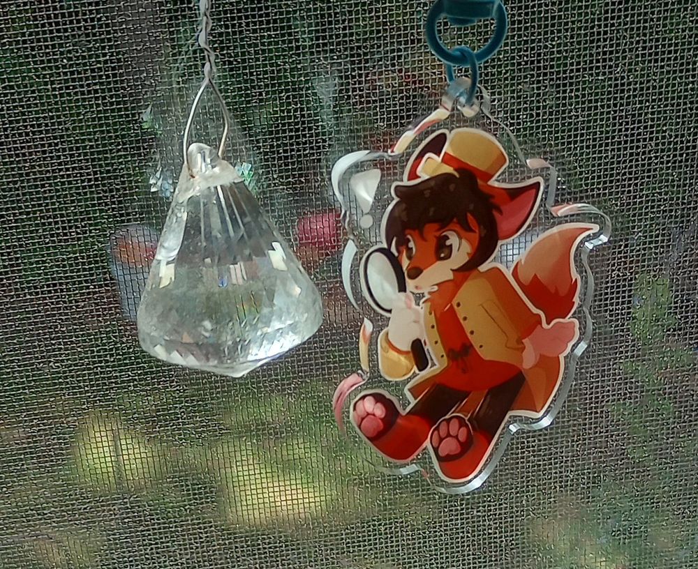 Keychain from the previous slide [A small anthropomorphic fox with a tan coat, red dress/shirt underneath, and black pants. Also wearing a small tan tophat with a red stripe, they are looking into a large magnifying glass. An inquisitive expression enhanced by an exclamation point above their head.] being held up next to a window decoration that looks like an upside down diamond. 