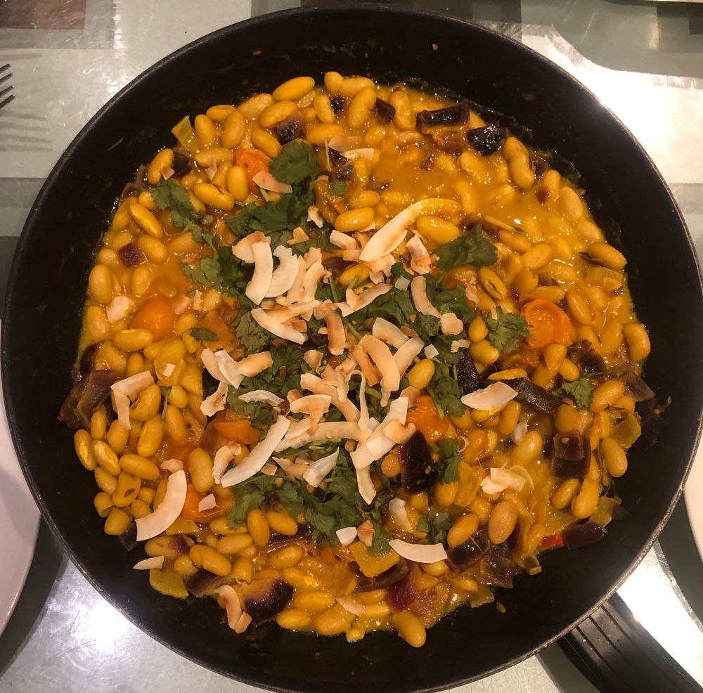 A black pan contains cannellini beans, sliced baby orange tomatoes, chopped onion - on top, toasted coconut flakes & coriander
