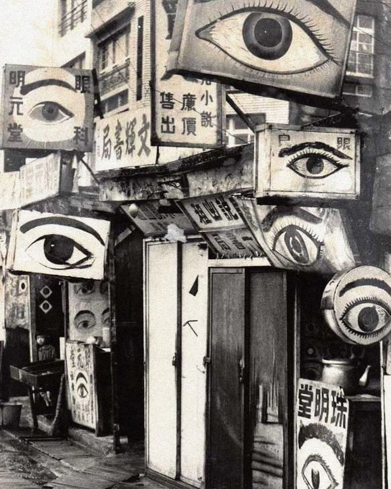 Foto antiga em preto e branco de entrada consultório de oftalmologia no Japão com muitos banners e placas na frente, alguns com caracteres japoneses mas vários com desenho de olho (1 olho por cartaz), criando uma vibe meio creepy/psicodélica