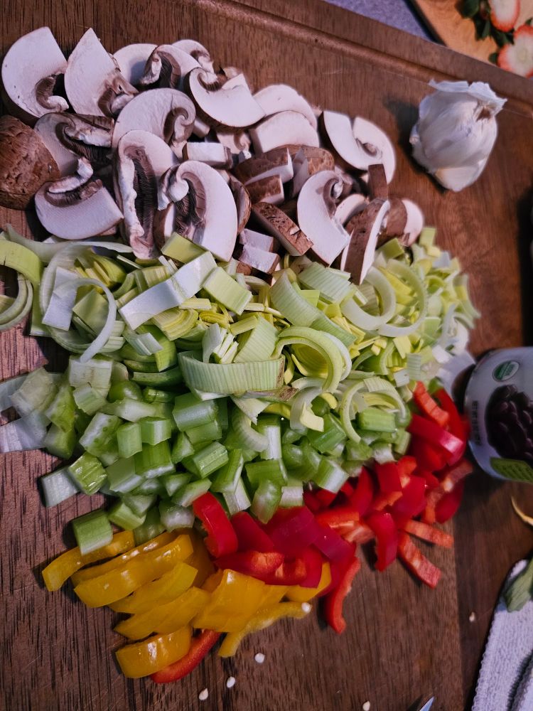 A cutting board with chopped mushrooms, leeks, celery, and yellow and red peppers.