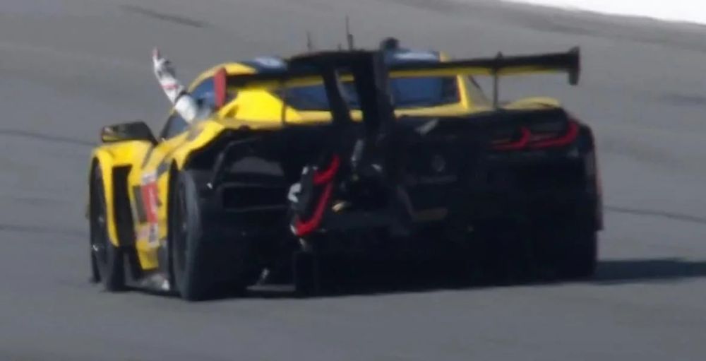 A bright yellow racing Corvette with damage to the rear of the car. The driver is sticking his arm out the small window, flipping the bird to another car.