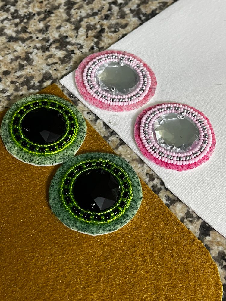 For beaded earrings in the price of the process of being made, there are two that are black and green, and two that are pink and silver. They are on backing. The backing is brown on the underside green on the regular side and white on the inside and pink on the outside.