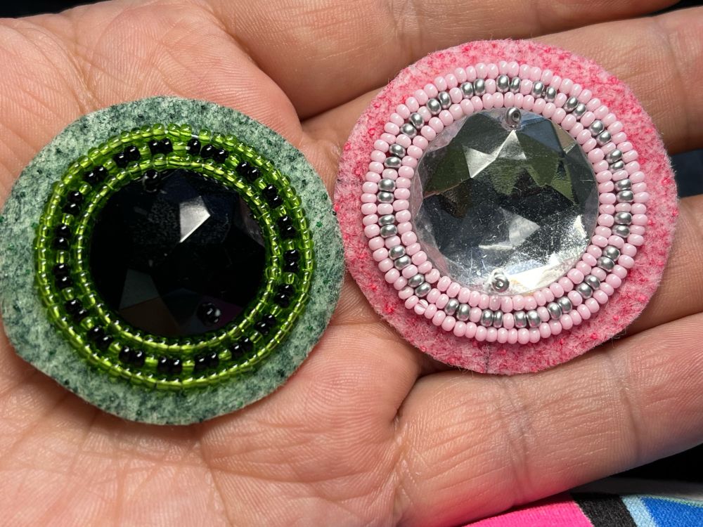 Parts of a beaded earring in my hand one has a black centre with green and black beads around it. The other has a silver centre with silver and pink beads around it