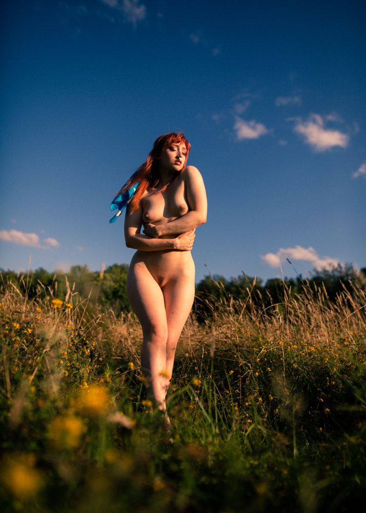 Nude model with her arms wrapped around her midsection standing in a field of grass against a blue sky with sparse clouds at golden hour