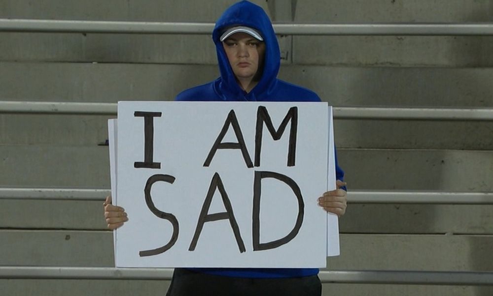 A person in a blue hoodie is sitting on bleachers, holding a large sign that says "I AM SAD."