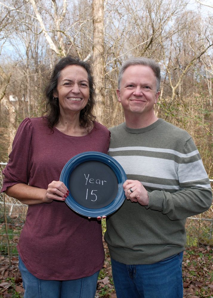 A middle-aged white couple smiling for a portrait. They're holding a plate between them that has "Year 15" chalked onto the surface.