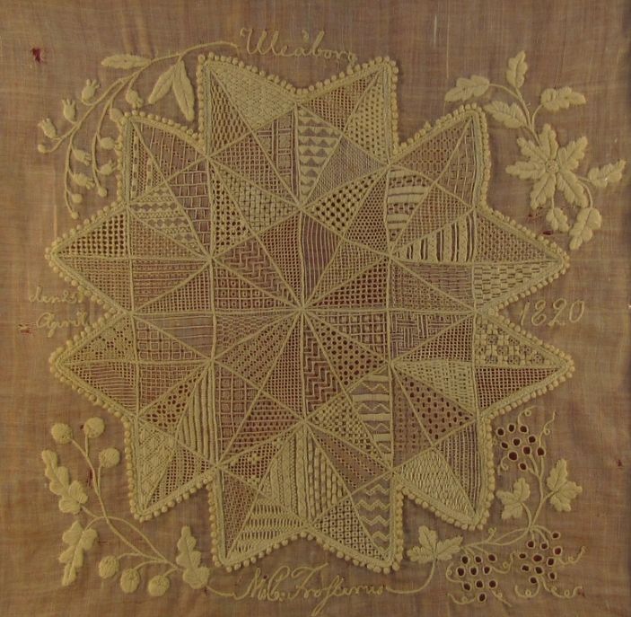 Photograph of a piece white on white embroidery in the shape of a star with flowers and vines around the edges; the star is divided into many smaller triangles each with its own stitch pattern.