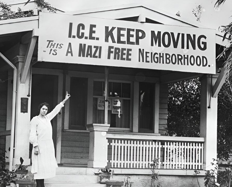   
A black and white photo features a woman standing on the steps of her house, pointing at a prominent sign above her porch that boldly asserts, "I.C.E KEEP MOVING This is a Nazi Free Neighborhood." Surrounded by plants and framed by a wooden porch and picket fence, the image conveys a strong anti-fascist message, combining historical context with modern political activism. The identity of the woman is not disclosed, but her stance reflects a commitment to social justice.  
